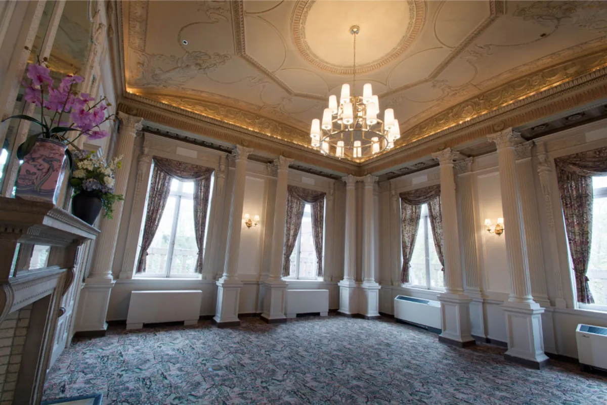 A luxurious room adorned with a glittering chandelier, ornate ceiling, expansive windows, and lush floral arrangements. The setting is designed for upscale gatherings, wedding receptions, and refined dinners, capturing the essence of romance and sophistication.