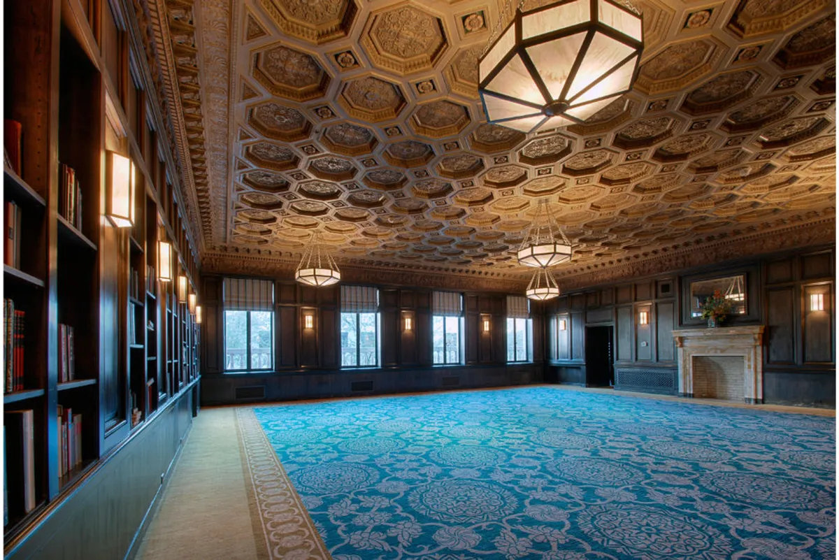 This elegantly appointed space features a sophisticated blend of ornate ceiling details, a stately fireplace, large windows bringing in natural light, and bookshelves that hint at intimate moments and quiet conversations. The decorative carpet and arrangement evoke an inviting yet luxurious atmosphere.