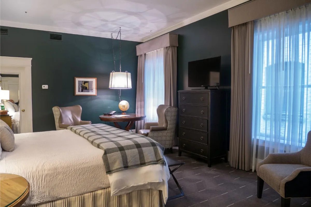 The image showcases a serene hotel room interior featuring a plush king-sized bed, inviting armchairs upholstered in leather and tweed, a handsome wooden dresser, and a softly glowing table lamp. The seating nook is designed for ultimate comfort, perfect for enjoying a quiet morning coffee or unwinding after exploring Portland's vibrant West End.