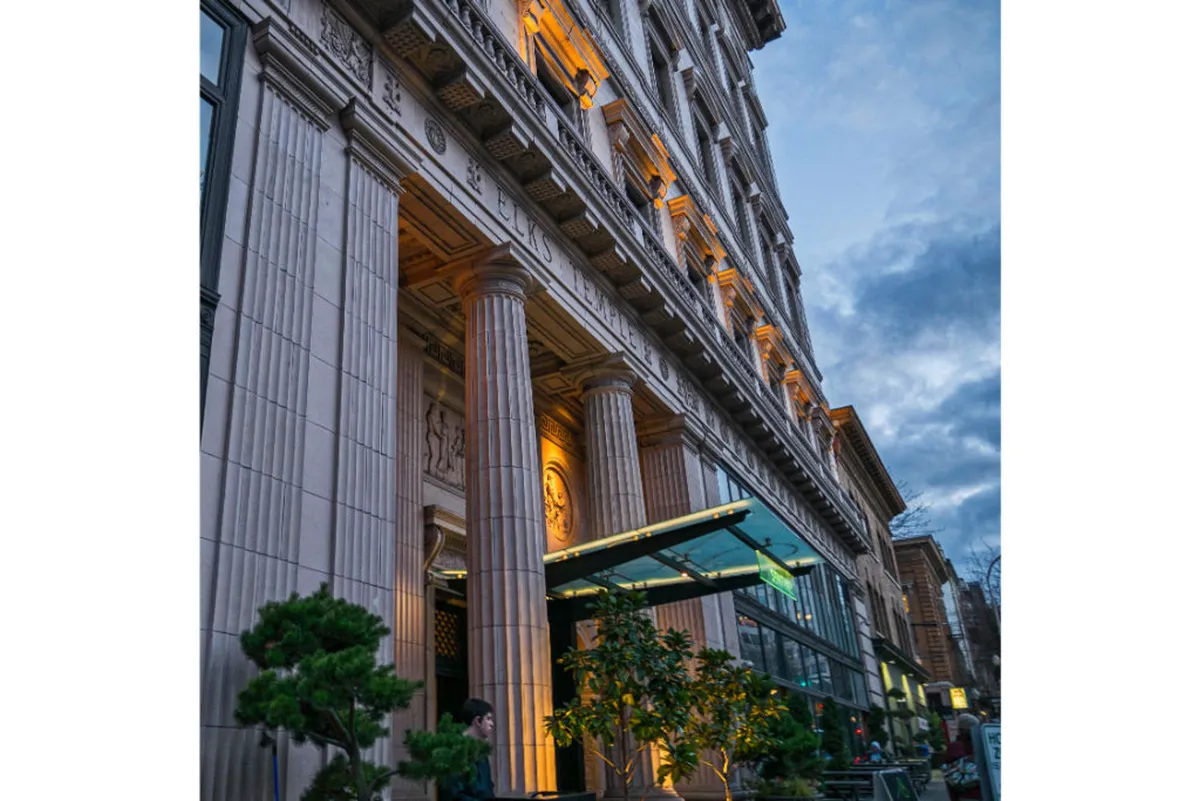 An inviting exterior scene with ornate columns, an illuminated facade, a glass entrance, and manicured landscaping. The image evokes the ambiance of evening activity—dining outdoors, strolling nearby, and enjoying the vibrant neighborhood.