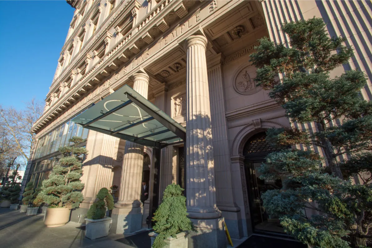 This exterior image showcases the hotel's grand entrance featuring stately columns, a glass canopy, and manicured greenery, with guests being welcomed outside. The elegant and historic facade conveys a sense of arrival and luxury, setting the tone for a seamless and upscale stay.