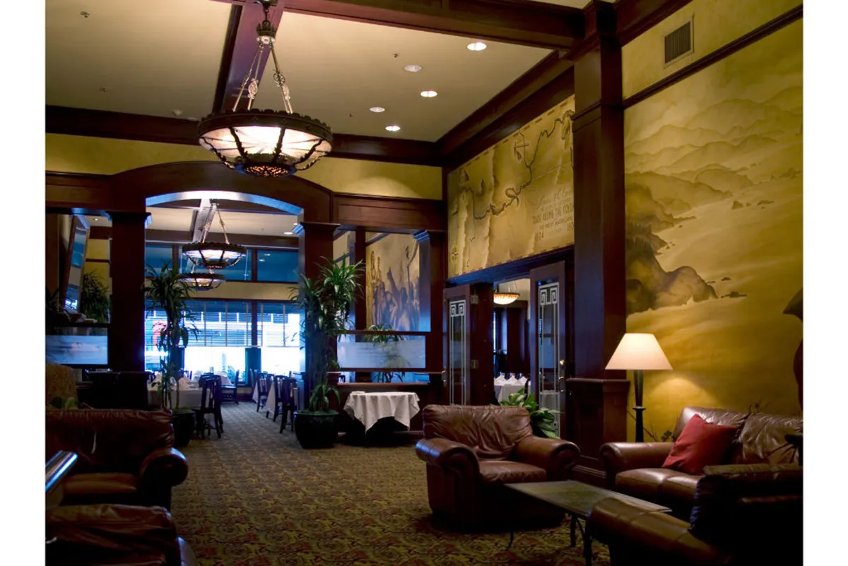 An elegant lobby area furnished with leather sofas, chandeliers, a decorative wall mural, and dining tables. The scene exudes both coziness and sophistication, creating a welcoming environment for arrivals, relaxing, and socializing.