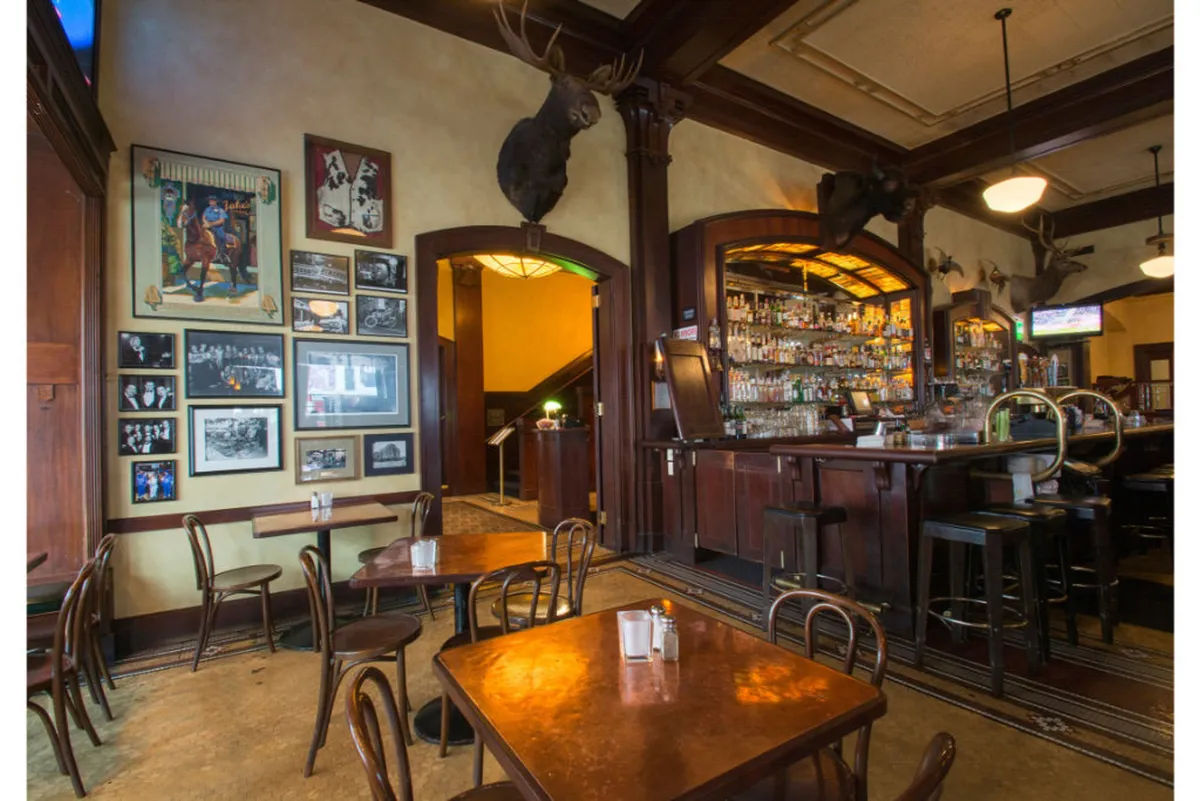A warm and inviting bar area featuring a polished counter, eclectic wall decor that includes photographs and mounted moose heads, and clusters of tables and chairs arranged for easy socializing. The space has a casual, welcoming energy, ideal for gathering with friends or unwinding after a day exploring Portland. The lighting and atmosphere evoke comfort and friendliness.